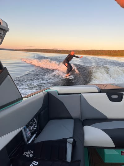 In Gromovo, Russia, a person in a black wetsuit surfs on a wake behind a boat at sunset. The surfer is on a white board, carving a wave. The boat, viewed from the stern, has gray and white upholstered seating. The water is calm with a large wake behind the boat. A line of trees borders the horizon, and the sky is a gradient of pale blue and orange. A black life vest is visible on the floor of the boat.