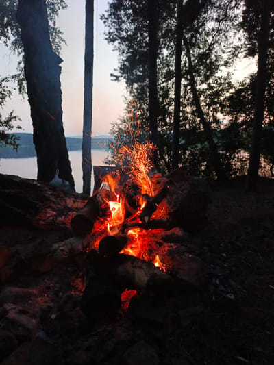 A campfire burns brightly in a forest clearing near a body of water, likely a lake or river, as evidenced by the gentle ripples and shoreline visible in the background. The setting appears to be Nepryakhinskoye Sel'skoye Poseleniye, Russia, given the context. The fire is composed of several logs, with bright orange flames licking upwards and a shower of orange sparks erupting into the dusky sky. The surrounding area is dark, with silhouetted trees forming a dense canopy overhead. A large, dark tree trunk is prominent on the left side of the frame, its bark rough and textured. In the mid-ground, to the left of the fire, a metal object, possibly a pot or kettle, hangs from a branch or support. The ground is covered in rocks and the charred remains of previous fires, with patches of dry leaves and twigs. The overall mood is one of a peaceful, rustic evening in nature. There are no people visible in the image.