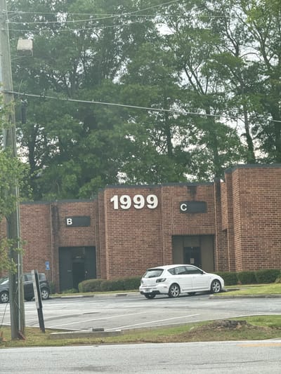 The image shows a brick building located at 1999, identified by large white numbers on the brick facade. The building has sections labeled "B" and "C". A white car is parked on the paved area adjacent to the building. The location is in Mountain Park, United States of America. A wooded area with tall trees is visible in the background. Overhead power lines are present. Another vehicle is partially visible at the bottom of the image. The scene appears to be on a cloudy day.