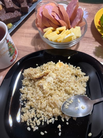 The image shows a close-up of a black plate filled with bulgur wheat, likely a meal. A silver spoon rests on the edge of the plate, with some bulgur grains on it. To the left, a white mug with a Christmas tree design is partially visible, containing a dark liquid, likely tea. In the background, a glass bowl contains sliced sausage and cheese. Behind that, a gray rectangular dish holds several pieces of dark chocolate. The scene appears to be set on a wooden table. The image does not provide any specific information to determine its location in Bely Klyuch, Russia.