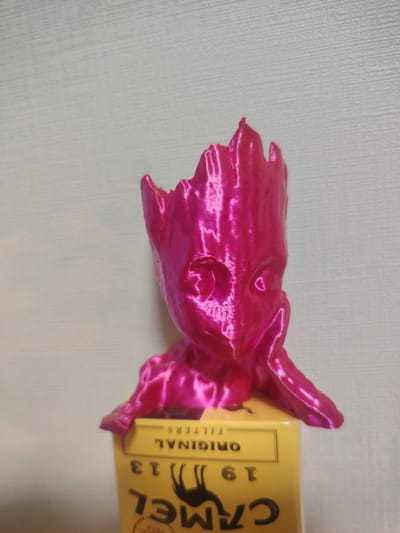 A bright pink, 3D-printed figurine resembling the character Groot from Guardians of the Galaxy is placed on top of a yellow cigarette pack. The figurine is small, with a detailed face and jagged head ornamentation. The cigarette pack, with the brand name "CAMEL" visible and printed in black text, appears to be a promotional item. The background is a light gray, textured wall. The scene is indoors, and the lighting is even, suggesting it was taken in a home or office environment. There is no indication of the location in Сухарево, Russia.