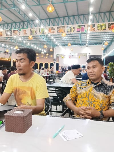 The image depicts an indoor scene within a casual dining establishment in Namplohbaro, Indonesia. Two adult males are seated at a white table in the foreground. The male on the left, of Asian ethnicity, wears a yellow short-sleeved t-shirt with a dark graphic and has dark hair with some facial hair; he is looking downwards and to his right. The male on the right, also of Asian ethnicity, wears a long-sleeved yellow shirt with a black and white intricate batik pattern; his hands are clasped together on the table, and he wears a dark watch or wristband on his left wrist, looking slightly to his left.
On the white table in front of them, a brown, woven-textured tissue box, a light blue pen, and a white paper receipt or order slip are visible.
The background shows a spacious area with a high ceiling supported by metal trusses. Numerous string lights and woven pendant lamps illuminate the space. A banner displaying various beverage and food images hangs horizontally along an upper wall section. Multiple other patrons, including men and women (some wearing headscarves), are seated at dark tables and chairs throughout the establishment. Arched decorative structures with lights are visible further back, and a large fan is mounted near the ceiling. The environment appears brightly lit by artificial light, suggesting evening or nighttime.