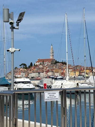 The image depicts a waterfront scene in Grad Rovinj, Croatia, with a view of the city's distinctive skyline, dominated by the bell tower of the Church of St. Euphemia. In the foreground, there's a metal railing with a sign indicating that access is restricted to crew only, written in multiple languages. A surveillance camera and streetlight are visible on a metal pole to the left. Several sailboats are docked along the waterfront, with their masts extending high into the blue sky. The buildings of Rovinj are clustered together, creating a dense and colorful cityscape with a mix of terracotta roofs. The blue water of the harbor can be seen in the background, with reflections adding depth to the scene.