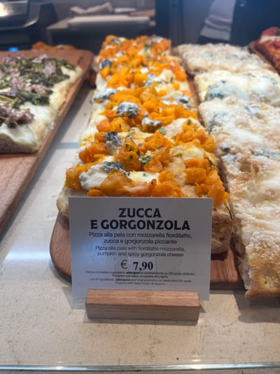 This media file captures a close-up view of various slices of pizza alla pala displayed in what appears to be a food counter, likely a pizzeria or bakery, in Dorno, Italy. The main subject is a rectangular slice of pizza topped generously with diced orange pumpkin (zucca), chunks of blue-veined gorgonzola cheese, and finely chopped green herbs, all resting on a creamy white base and focaccia-style dough.
Behind this prominent slice, another pizza with a lighter, perhaps cheesy or herb-based topping is partially visible. Further in the background, another pizza slice with darker, possibly vegetable or meat toppings, can be discerned, though it is out of focus. All pizzas are presented on light-colored wooden cutting boards, emphasizing a rustic and artisanal aesthetic. The setting suggests an indoor commercial environment with bright, possibly artificial, lighting illuminating the food.
A white price tag is strategically placed next to the pumpkin and gorgonzola pizza. The visible text on the tag reads:
* "ZUCCA E GORGONZOLA"
* "Pizza alla pala con mozzarella fiordilatte, zucca e gorgonzola piccante"
* "Pizza alla pala with fiordilatte mozzarella, pumpkin and spicy gorgonzola cheese"
* "€ 7,90"
* Smaller text at the bottom indicates: "Elenco completo ingredienti, allergeni e caratteristiche su QR code dedicato. Prodotto con base congelata all'origine. List of ingredients, allergens and characteristics on dedicated QR code. Product with base frozen at source."
The image depicts a selection of freshly prepared pizza slices, highlighting their ingredients and pricing for customers, consistent with a food service establishment in Italy. No people are visible in the frame.