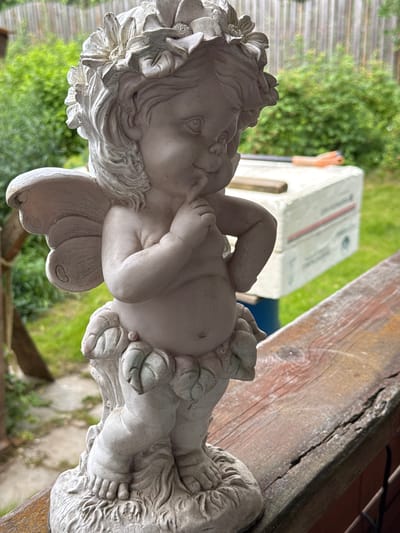 A light pink, weathered statue of a cherub or fairy stands on a weathered wooden railing. The statue is of a chubby child with delicate wings, adorned with a floral wreath on its head, and appears to be in a thoughtful pose with a finger to its chin. The background is blurred, showing greenery and a wooden fence, suggesting an outdoor setting, possibly a garden or backyard. In the middle ground, a white object, possibly a cooler or storage container, is visible with some tools resting on it. The scene appears to be located in Bersenevka, Russia.