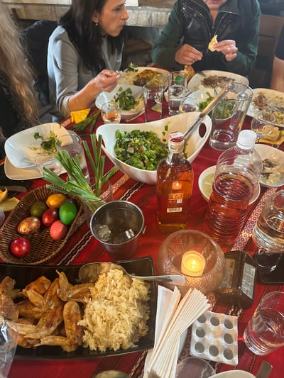 A horizontal shot depicts a bustling table scene, potentially a meal shared among people, situated in Sadievo, Bulgaria. The table is draped with a red tablecloth featuring a geometric pattern.

Around the table, people are seated, their arms and hands visible as they reach for food. One person, possibly female, with long brown hair and wearing a grey top, is visible on the left side of the frame. Opposite her, a person with short dark hair wearing a green jacket is holding a piece of food.

The table is laden with an assortment of food items. Several plates with salad are visible, and one contains a dish of meat. There is also a platter with rice and what appears to be fried chicken wings. Additionally, there is a basket filled with dyed eggs.

Multiple bottles and jars are placed on the table, along with drinking glasses. A candle holder with a lit candle adds a warm glow to the scene. A black smartphone and a white egg carton complete the scene.
