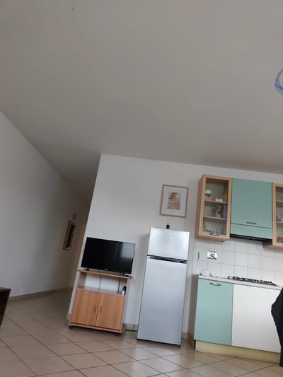 This is a medium-angle, eye-level, interior shot of a kitchen and adjacent living area in Guarene, Italy. The scene is bright and appears to be daytime, with natural light coming from an unseen source. The walls are painted white, and the floor is tiled with light brown squares.
In the living area, a light brown wooden TV stand with two cabinets sits against the wall. A black flat-screen television is placed on top of the stand. To the left of the TV stand, a dark brown piece of furniture, possibly a coffee table, is partially visible. Further down the wall, a framed picture with an abstract design of leaves and an orange shape is hanging. A rectangular mirror with a thin wooden frame is also mounted on the wall.
The kitchen area features a silver refrigerator on the left. Adjacent to it is a white tiled backsplash above a countertop. The kitchen cabinets are a mix of colors: a wooden cabinet with glass doors above the countertop, a light teal cabinet with a white door below the countertop, and a white cabinet next to that. A gas stovetop is visible on the countertop. The overall impression is that of a lived-in, functional space.
There are no people visible in the media file. The primary objects are the refrigerator, television, TV stand, kitchen cabinets, countertop, stovetop, framed picture, and mirror. The setting is a domestic interior, likely a kitchen and living room combination. No specific activity is taking place, suggesting a static shot of the environment. There is no visible text in the image.
