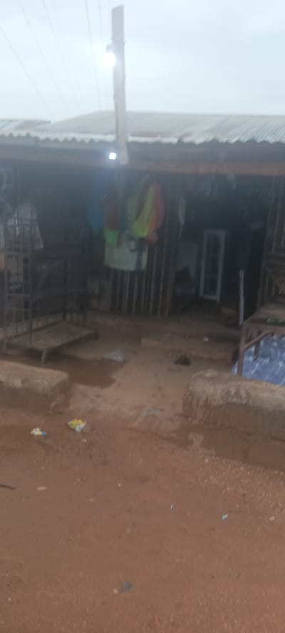 The image depicts a small, open-fronted shop in Ladura Foron, Nigeria. The shop is constructed with corrugated metal roofing and possibly metal or wooden vertical supports. Hanging within the shop are articles of clothing, including what appears to be a brightly colored safety vest. Inside, objects, including what may be a window frame, can be seen in the dim interior. The foreground consists of a dirt ground, littered with small pieces of debris and trash. Low concrete blocks define the edges of the shop's immediate surroundings. A utility pole with an attached light fixture is situated in front of the shop's structure.
