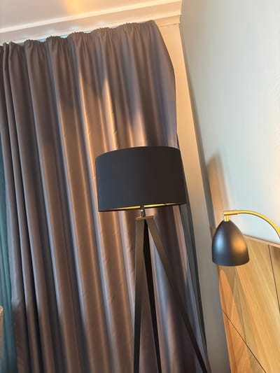 A modern, dark gray floor lamp with a black cylindrical shade stands in the foreground, angled slightly to the right, positioned in front of matching floor-to-ceiling curtains. The curtains, made of a lustrous fabric, are drawn partially open, revealing a sliver of a light blue curtain behind them. The room is dimly lit, with warm light spilling from an unseen source, casting highlights on the curtains and the lamp shade. To the right of the lamp, a portion of a headboard, possibly made of wood with a prominent grain pattern, is visible. A smaller, black lamp with a gold arm is perched on top of the headboard. The wall behind the headboard is painted a light, neutral color. The scene appears to be indoors, likely in a hotel room in Malmö, Sweden, given the context of the prompt. No people are present in the image.