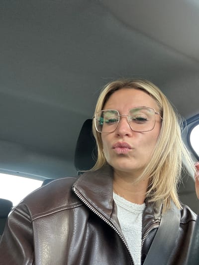 A female individual is depicted from the mid-chest up, seated inside a vehicle in Villeneuve-Loubet, France. She has blonde, shoulder-length hair and wears large, square-framed glasses with light-colored metal frames. Her lips are pursed in a puckering expression. She is clad in a dark brown, zippered jacket, possibly of faux leather, over a light-colored, textured top, and a black seatbelt crosses her upper torso. The vehicle's dark grey headliner and a black headrest are visible in the background. A bright, undifferentiated light is visible through a window on the right side of the frame.