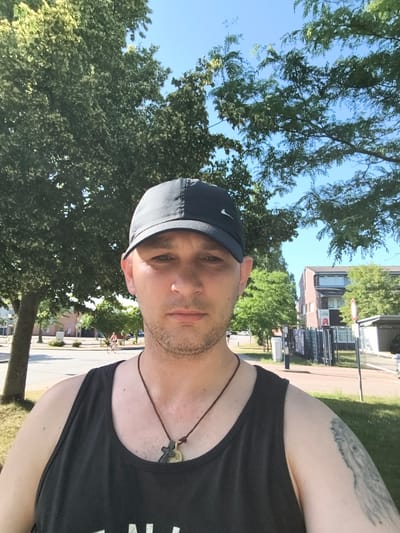In a selfie taken outdoors, a Caucasian male with short brown hair, stubble, and fair skin is looking directly at the camera. He is wearing a black tank top and a dark baseball cap. Around his neck, he has a brown cord with a pendant. A tattoo is visible on his right arm. The background consists of trees, buildings, and a street with a person riding a bicycle in the distance. The sky is clear and blue. The scene appears to be set in a public area, possibly a park or street in the city of Wolgast, Germany, given the presence of what appears to be a pharmacy sign with German text and a German red and white traffic sign.