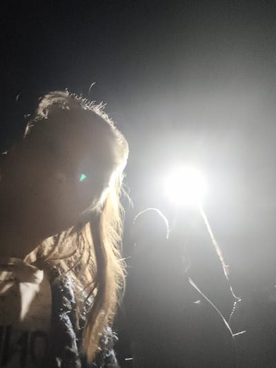 In Il'mino, Russia, an individual is captured in a close-up, side profile, illuminated by a bright light source. The person's face is partially obscured by shadow and the strong backlight, with only the outline of their head and flowing, light-colored hair visible. The hair appears to be long and somewhat disheveled, with individual strands catching the light. A faint green lens flare is present on the left side of the image, near the person's face. In the background, a second, darker silhouette of a person is faintly visible. A thin, vertical object, possibly a pole or stick, extends upwards towards the light source, with small, bead-like objects or attachments visible along its length. The overall environment is dark, suggesting nighttime or an indoor setting with limited ambient light.
