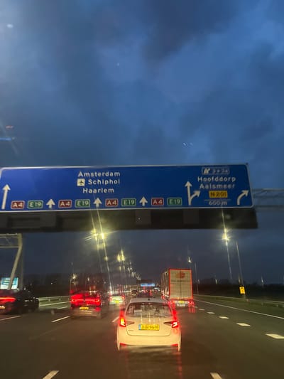 The image captures a nighttime or dusk scene on a multi-lane motorway in the Netherlands, specifically on the A4 approaching the exit for Hoofddorp and Aalsmeer. The road surface is wet, reflecting the yellow light from overhead streetlights and the red taillights of vehicles. A large blue overhead gantry sign is prominent, directing traffic straight ahead for Amsterdam, Schiphol, and Haarlem via the A4 and E19, and indicating a right turn in 600 meters for Hoofddorp and Aalsmeer via the N201 (exit 3+2a).

In the immediate foreground, from the perspective of a vehicle, a white Skoda Fabia sedan with Dutch license plate "JH-96-H" is visible, its taillights illuminated. Ahead of it, in the middle lane, is a dark-colored SUV, possibly a Volkswagen T-Roc, with Dutch license plate "Z-564-JF," also displaying illuminated taillights. To the right, in the adjacent lane, a white semi-trailer truck with "VAN DRIMMELEN" branding on its trailer is visible, its rear lights active. Numerous other vehicles with illuminated red taillights extend into the background, indicating active traffic. The sky is dark and cloudy. This precise location is on the A4 motorway, 600 meters prior to the Hoofddorp/Aalsmeer exit, within the city of Hoofddorp, Netherlands.