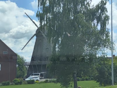The image depicts a traditional windmill, a classic example of historical architecture, situated in the picturesque locale of Ceraukste, Latvia. The windmill, with its imposing structure and wooden vanes, stands as a testament to a bygone era. It is surrounded by lush greenery, with trees and shrubs forming a verdant backdrop. In the foreground, a well-maintained lawn stretches out, suggesting a park-like setting. The sky above is a canvas of blue, dotted with fluffy white clouds, casting a gentle light on the scene. The presence of a white car and a white van indicates that the location is accessible and likely serves as a tourist attraction or a place of historical interest. The overall atmosphere is one of tranquility and natural beauty, inviting visitors to explore and immerse themselves in the charm of rural Latvia.
