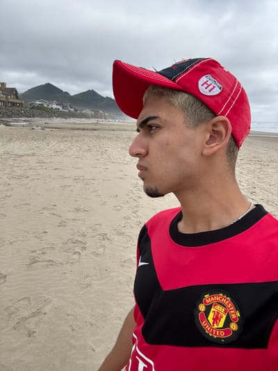 A person stands sideways on a beach. They wear a red and black Manchester United soccer jersey and a red baseball cap with white stitching and a “H1” patch. The person has short, light-colored hair and a trimmed goatee. The beach stretches into the distance with a cloudy sky above. Behind the beach is a view of mountains and houses. The sand shows footprints and subtle patterns.