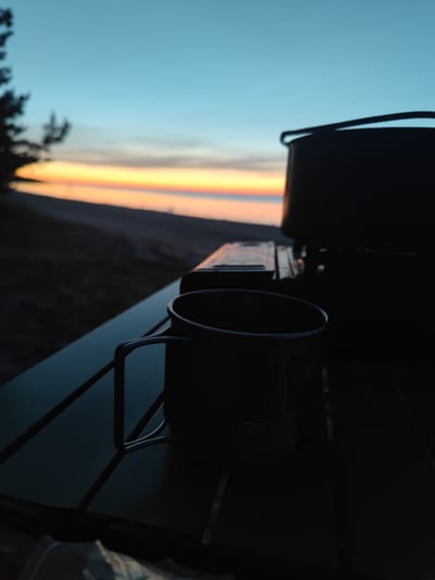A silhouette of a camping mug and a piece of camping equipment sits on a dark, folded picnic table. In the background, the sky transitions from a deep blue to hues of orange and yellow, indicating either sunrise or sunset over a body of water. The shore is visible, with dark foliage on the left side of the frame. The location is identified as Motornoe, Russia.