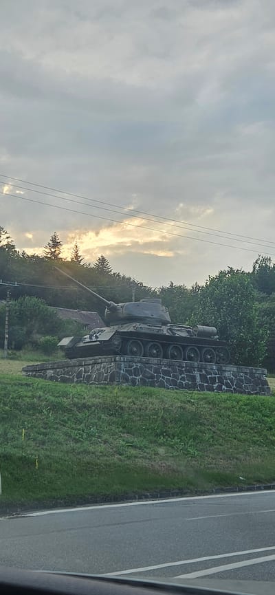 The image shows a monument consisting of a Soviet-era T-34 tank on a stone-covered pedestal, located in the village of Vyšný Komárnik, Slovakia, near the Dukla Pass. The tank is positioned on a grassy slope with trees in the background. The sky is overcast with patches of sunlight breaking through the clouds, suggesting it is either sunrise or sunset. A road is visible in the foreground, indicating the monument is roadside. There are power lines overhead. No people are present in the image.