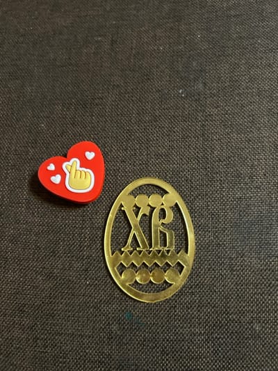 The scene depicts two distinct small objects positioned on a dark brown, finely woven fabric surface in Dmitriyevskaya, Russia. The fabric displays a uniform texture with minimal lint and a faint bluish discoloration in the lower right quadrant. To the upper left, a red, heart-shaped charm with a white border measures approximately 2.5 cm at its widest point. It features a yellow hand gesture, commonly known as a "finger heart," outlined in white, and three smaller white heart shapes surrounding it. To the lower right, a golden-hued, flat, oval-shaped openwork ornament, approximately 4 cm in height, resembles an Easter egg. Within its design, the Cyrillic letters "Х" and "Б" are prominently displayed above a decorative band of repeated jagged peaks, which in turn sits above a row of five small circular cutouts. Both objects are stationary and appear to be charms or decorative items.