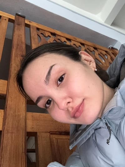 A close-up selfie depicts a young adult female indoors in Parizhskaya Kommuna, Russia. The subject's head is tilted, and she is looking directly at the camera with dark eyes. Her dark hair is visible, and her complexion is fair. She is wearing a light blue or grey puffer jacket; a black, beaded zipper pull is visible on the front. Her visible right ear features a clear stud earring in the earlobe and a small metal ring on the helix. The background includes a light brown wooden structure, possibly a bed frame or room divider, characterized by vertical slats and an ornate carved design along its upper section. A plain, light-colored wall or ceiling is visible above the wooden structure.