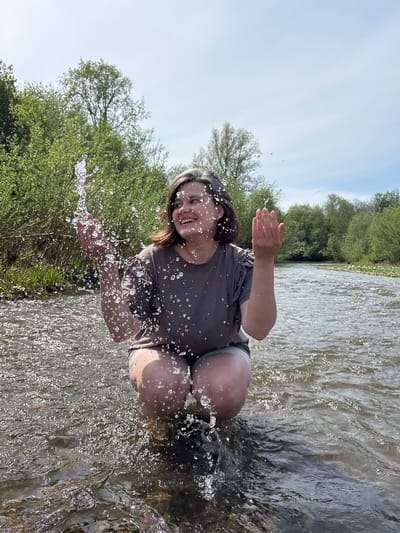 A woman is squatting in a river, splashing water with her hands. The background consists of trees and the sky. The location is Усть-Тунтор, Russia.