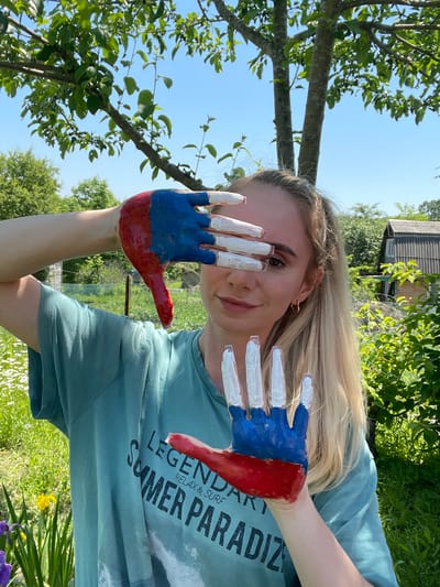 A young Caucasian woman with blonde hair pulled into a ponytail stands outdoors, partially obscuring her face with her hands, which are painted with red, white, and blue. She is wearing a teal t-shirt with the words "LEGENDARY SUMMER PARADIZE RELAX & SURF" printed on it. Her right hand, painted in red and blue, is held up to her face. Her left hand, painted white on the fingers and blue and red on the palm, is held in front of her. The background shows lush green trees, foliage, a wooden fence, and a rustic wooden building, suggesting a rural or suburban setting. The sky is clear and blue. The overall lighting indicates daytime. This scene is likely located in the city of Yasnoe, Russia, given the context of the provided information.