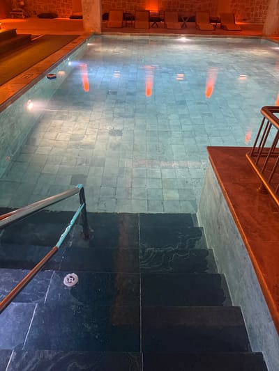 The image shows an indoor swimming pool with glass walls, likely located in a hotel or spa in Ürgüp, Turkey, as the architecture and lighting suggest a luxury environment commonly found in the region's cave hotels.
The pool itself has a tiled bottom with embedded lights creating reflections. Adjacent to the pool are dark-colored stone stairs leading downwards. A metal railing is visible alongside the stairs and at the edge of the pool. A series of chairs or seating is located on an elevated ledge along one side of the pool, illuminated with warm lighting. The pool's water is still, mirroring the surrounding environment.