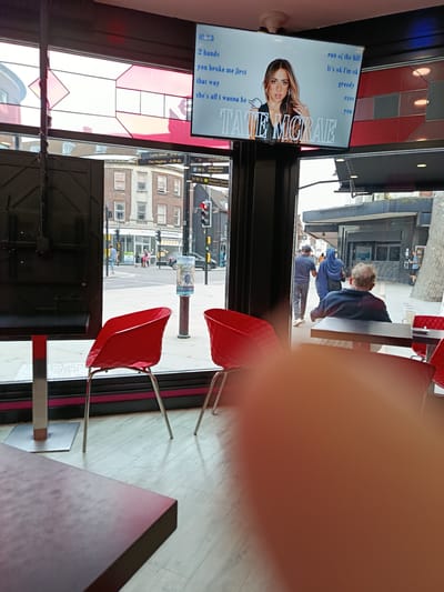 The image shows an indoor scene, likely a cafe or restaurant, with a large screen displaying a music video for Tate McRae. The screen shows the artist's name "TATE MCRAE" in large font, along with song lyrics such as "2 hands", "you broke me first", "that way", "she's all i wanna be", "run of the hill", "It's ok I'm ok", "greedy", "exes", and "you". In the foreground, two bright red chairs are visible, along with a table partially obscured by a blurred orange area, suggesting the photographer's hand.
Through the window, a street scene in Bedford, United Kingdom, is visible. Signposts indicate "Lurke Street", "The Higgins" (a museum and art gallery), and "Church Arcade". The architecture suggests a typical British town center. People are walking on the street, and traffic lights are visible. One signpost indicates "Harpur Centre" and "Howard Centre". A red traffic light is illuminated. The environment appears to be a public space with commercial establishments.