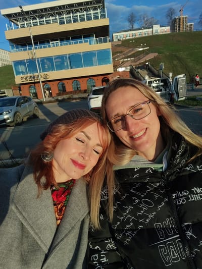 Two women pose closely together for a selfie in the city of Средний Постол, Russia. The woman on the left has red hair, wears a gray coat over a colorful scarf, and a gray beanie. Her eyes are closed and she wears a large, decorative earring. The woman on the right has long blonde hair and wears glasses and a black jacket with white lettering. Both women are smiling.
In the background, a modern building with large windows sits adjacent to a parking lot containing multiple cars. Behind the building, a grassy hill leads to a series of walkways, stairs, and buildings. A small vehicle with an open side panel, possibly a coffee or food stand, is visible on the walkway. A construction crane is visible in the distance. The sky is blue with some clouds.