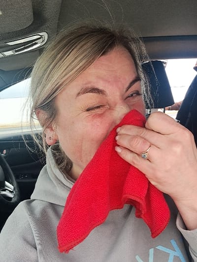An adult female, with light-colored hair and visible facial redness around the nose and cheek, is depicted inside a vehicle, specifically in the city of Sasovo, Russia. She is wearing a light gray hooded sweatshirt and holds a folded red microfiber cloth against her nose and mouth area with her right hand. Her right eye is partially visible and squinted, while her left eye is closed, indicating a strong sensory reaction, possibly a sneeze or irritation. A gold-toned ring with a light-colored gemstone is visible on her right ring finger. The vehicle's interior features a gray headliner, an overhead light fixture, and part of a black steering wheel on the left. A dark-clad figure or object is partially visible through a window in the right background.