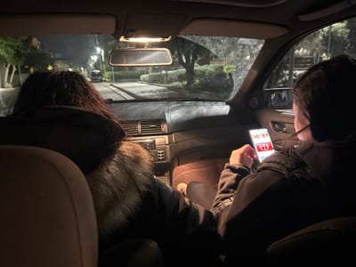 The image depicts the interior of a vehicle at night, viewed from the rear passenger compartment, situated in Glyka Nera Municipal Unit, Greece. Two individuals are present in the front seats. The occupant of the driver's seat, on the left, has dark, possibly curly, hair and wears a dark jacket with a light-colored fur or faux-fur trimmed collar, facing forward. The occupant of the front passenger seat, on the right, wears a dark padded jacket and holds a white smartphone with their left hand, actively viewing its brightly illuminated screen which displays content featuring red and white elements. The vehicle's dashboard, central air vents, and stereo controls are visible. An overhead interior light is active, casting light downwards. Through the front windshield, a dimly lit urban or suburban street is discernible, with outlines of trees, buildings, and the distant lights of another vehicle.