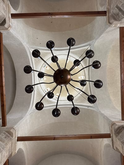 The image shows a view looking up at a large chandelier suspended from the arched, white-painted ceiling of a building. The chandelier has a dark bronze or black finish and features a central sphere with numerous arms extending outward, each terminating in a rounded cup-like fixture. Light bulbs are visible within some of these cups. The ceiling is divided into several arched sections by a series of wooden beams that run along the edges of these sections. The walls are constructed of stone. The photograph likely was taken within Manasija Monastery, Despotovac Municipality, Serbia.