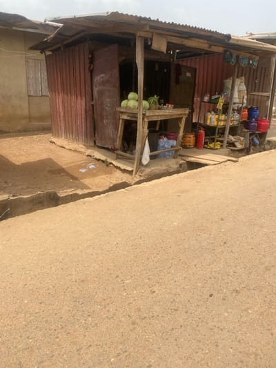 An outdoor scene in Odo Ona, Nigeria, depicts a roadside commercial establishment under daylight conditions. The foreground features a light brown asphalt road with visible aggregate. A concrete gutter, approximately 30-40 cm wide, runs parallel to the road, separating it from a dirt and concrete shoulder. To the left, several pieces of discarded paper or debris are visible on the dirt. A small, rustic shop constructed from reddish-brown corrugated metal sheeting and rough-hewn wooden posts and beams occupies the central-right. It features an open facade with a corrugated metal roof.
On the left side of the shop's opening, a wooden display table holds six large green watermelons and one pineapple, along with a small white dish containing cut fruit. Beneath this table, a stack of approximately ten clear plastic bottles of water (likely 19-liter capacity) rests on the ground, and a white cloth hangs from the table's support. Further into the shop, to the right, a multi-tiered display rack holds various items: multiple yellow plastic containers (possibly motor oil), a smaller rack with assorted colorful plastic containers (red, orange, purple, blue, green), two red fire extinguishers, and two orange gas cylinders. Above this area, several pairs of colorful flip-flops or sandals are visible hanging. A wooden plank serves as a bridge over the gutter on the right side of the shop, providing access from the road. To the immediate left of the shop, a simple building with a light-colored, faded wall and a boarded-up window is partially visible. No individuals are present or engaged in activity within the frame.