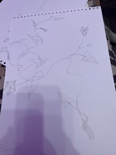 The image shows a pencil sketch of a vine with flowers and leaves on a white sheet of paper. The sketch is done in a delicate, detailed style. The vine is shown in different stages of growth, with some buds and some fully bloomed flowers. The flowers are depicted with intricate petals, and the leaves are drawn with fine veins. The sketch is placed on a surface that appears to be a table with a patterned cloth. There is also a hint of another piece of paper in the background, also with a drawing. The bottom left corner of the paper has some handwritten Cyrillic text, which appears to read "Выонок" (Vy-o-nok), potentially a name or a title. The overall impression is of an artistic endeavor, a work in progress. The request to include the location in the city of Zmeinyi, Russia is not possible to fulfill as there are no geographical indicators in the image.