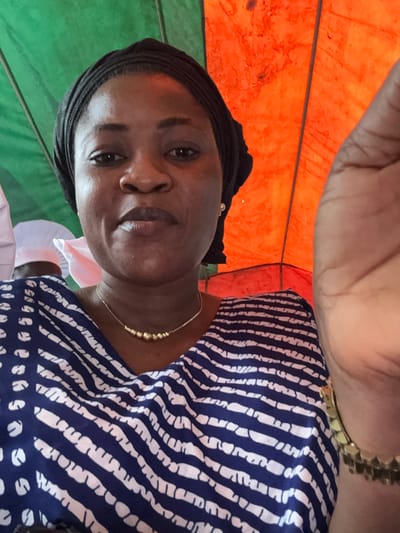 A woman is posing for a selfie in what appears to be an outdoor setting, possibly under a canopy or tent, with a green and orange backdrop. She is wearing a black headscarf and a blue and white striped top. A gold necklace with small beaded pendants adorns her neck, and a similar gold bracelet is visible on her right wrist. Her right hand is partially in the frame, obscuring part of the background. The woman's skin tone is dark, and she is looking directly at the camera. There are other individuals in the background, one wearing a white head covering. The lighting suggests it is daytime.