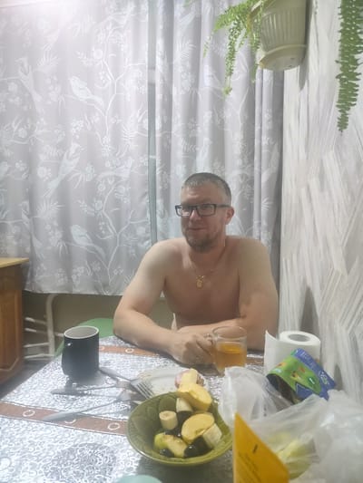 A man with short hair, glasses, and a goatee is sitting at a table, shirtless. He is wearing a gold chain with a pendant. In front of him on the table are a black mug, a glass of amber liquid, a plate with some food (likely cheese and ham), a bowl of fruit (including apples and bananas), a roll of toilet paper, and a plastic bag with some items. The man appears to be in the process of eating or having a meal. The setting appears to be a room with curtains that have a bird and floral pattern, and a textured wallpaper on the right side. A potted plant with green leaves hangs from the top right. The lighting suggests it could be daytime, possibly late afternoon or evening, given the indoor setting. The overall mood seems relaxed and casual.