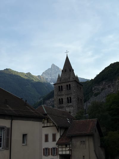 The image shows a view of the Saint-Maurice Abbey in Switzerland. The abbey, built with grey stone, features a tall, pointed spire topped with a cross. The architecture suggests a historical religious site. In the foreground, various buildings with tiled roofs are visible, some with arched windows and shutters, contributing to the rustic charm of the scene. Behind the abbey, a steep, green mountainside rises sharply, partially shrouded by trees. A rocky peak, possibly snow-capped, emerges in the distance, hinting at the alpine environment. The sky is overcast with a pale, diffused light, suggesting it is either early morning or late afternoon, or a generally cloudy day. The overall impression is one of tranquility and historical significance, set against a dramatic natural landscape. There is no visible text in the image.
