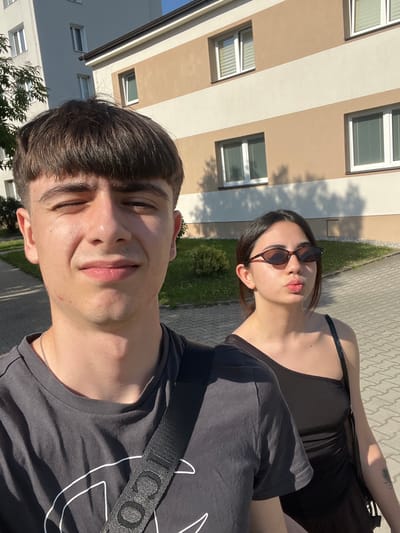The image is a selfie taken outdoors in Dobřany, Czechia. Two young adults are visible. In the foreground, a young man is looking at the camera with a slight smile. He has short brown hair and is wearing a dark t-shirt with a white graphic design and a silver necklace. Behind him and to the right, a young woman is also looking at the camera, with her head tilted back and her lips puckered. She is wearing sunglasses and a black top, with a tattoo visible on her arm. The background shows a building with beige and white stripes and several windows. The ground is paved with cobblestones, and there is some greenery visible. The lighting suggests it is daytime, likely late afternoon due to the long shadows. The overall mood appears casual and sunny. There is visible text on the strap of a bag worn by the man, which reads "ICONE".