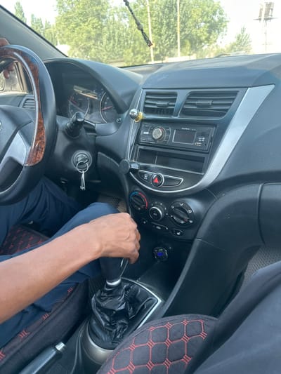 The image shows the interior of a car. A person's right arm and hand are visible, gripping the gear shift. The driver is wearing dark blue pants. The dashboard features a car stereo, air vents, climate control knobs, and a car key in the ignition. A USB car charger is plugged into a power outlet, displaying a blue light. The gear shift is covered in black plastic. The car seat has a black cover with a red honeycomb pattern. Outside the car, trees and bright daylight suggest it is daytime and likely clear weather. The scene appears to be set in a car, possibly on the road, given the driver's hand on the gear shift. The visible text on the stereo reads "SUPRA". The location is identified as Ынтымак, Kyrgyzstan.