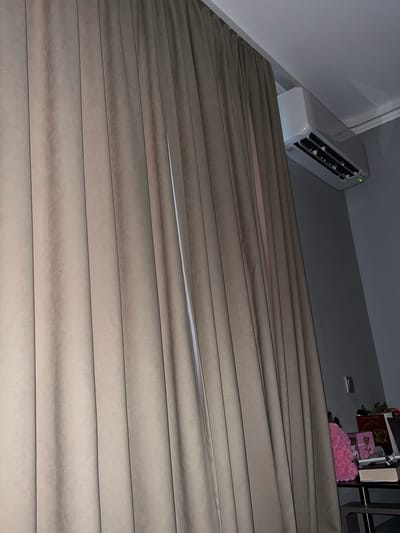 The image shows a close-up view of beige pleated blinds. An air conditioning unit is visible mounted on the wall near the top left. On a shelf behind the blinds (top right), there are books, a pink floral arrangement, and some decorative items. The scene is dimly lit. The specific location in Kribul, Bulgaria cannot be determined from the image. No people are visible.