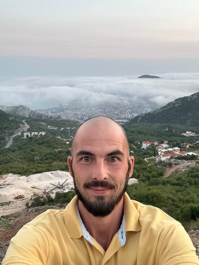 A medium shot selfie shows a bald, bearded man with light skin, dark eyebrows, and hazel eyes, wearing a yellow polo shirt with a blue and white striped collar. He is looking directly at the camera with a slight smile. In the background, a dense fog blankets a city, with only the tops of buildings and a distant hill with a communication tower visible. The fog extends to the horizon, where a pale sky meets the white haze. Below the fog, a lush green mountainside is dotted with red-roofed houses and a winding road. A construction site with piles of sand, heavy machinery, and trucks is visible in the foreground, to the left of the man. The man is positioned in front of a rocky, elevated terrain. The scene is consistent with an elevated viewpoint overlooking Markovići, Montenegro, during foggy weather.