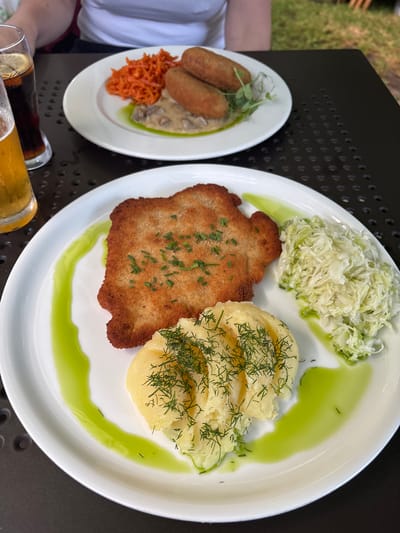 A close-up shot shows a white plate with a meal on a dark, textured outdoor table. The plate contains a breaded schnitzel topped with chopped green herbs, a serving of mashed potatoes garnished with dill, and a pile of shredded coleslaw. A vibrant green sauce is artfully drizzled around the food. In the background, another white plate with three croquettes, shredded carrots, and a creamy sauce is visible, alongside a glass of dark soda with ice and lemon, and a partially visible glass of beer. The setting appears to be outdoors, likely a restaurant patio in Bialowieza, Poland, with glimpses of grass in the upper right corner.