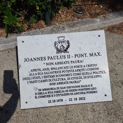A white marble commemorative plaque, rectangular in shape, lies horizontally on a light grey paved surface. The plaque features a black-inked coat of arms at the top center, above black capitalized text in Italian. The text reads "IOANNES PAULUS II - PONT. MAX." followed by his famous quote, "NON ABBIATE PAURA! APRITE, ANZI, SPALANCATE LE PORTE A CRISTO! ALLA SUA SALVATRICE POTESTÀ APRITE I CONFINI DEGLI STATI, I SISTEMI ECONOMICI COME QUELLI POLITICI, I VASTI CAMPI DI CULTURA, DI CIVILTÀ, DI SVILUPPO. NON ABBIATE PAURA!". Below this, an inscription states "IN MEMORIA DI SAN GIOVANNI PAOLO II E DELLA SUA OMELIA DI INIZIO PONTIFICATO IL COMUNE E I CITTADINI DI ANCONA POSERO", with dates "22 10 1978 - 22 10 2022". The plaque is secured by four visible screws or bolts, one in each corner. In the background, behind the plaque, green foliage and disturbed soil are visible, along with a dark stake and a small circular ground light. A dark, distinct shadow of a person holding a smartphone or camera is cast onto the lower left portion of the plaque and the ground. The scene is located in Ancona, Italy, commemorating Pope John Paul II and the 44th anniversary of his pontificate.
