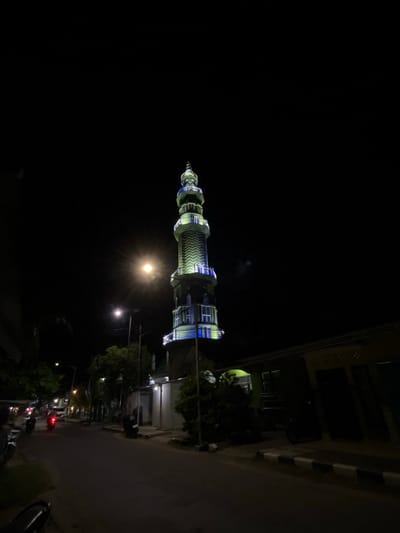 The media file shows a minaret illuminated by artificial lights at night. The minaret is tall and slender, with a conical top and intricate decorative patterns along its structure. Blue and yellow lights accent the minaret, making it stand out against the dark sky. The image is taken from a low angle, emphasizing the height of the minaret.
In the background, a building with windows is visible, also illuminated by lights. Trees and streetlights line the road, casting a warm glow on the surroundings. Several motorcycles are parked or being ridden on the road, suggesting a public or residential area. The overall scene is urban and lively, despite the darkness of the night. The image seems to be taken in Karimunjawa, Indonesia, based on the provided context.
The time of day is clearly night, as indicated by the dark sky and artificial lighting. The weather appears clear, with no visible rain or fog. The mood of the image is serene and perhaps even a little mystical, due to the dramatic lighting of the minaret. There is no visible text in the image. The media file captures a moment of nighttime beauty in an urban setting, highlighting a significant architectural structure.