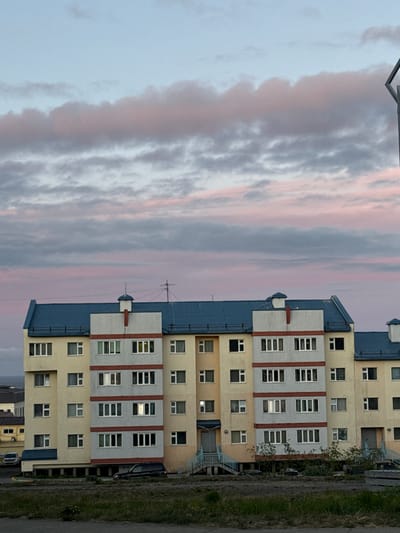 The image shows a multi-story apartment building with a blue roof, yellow and gray facade, and red trim. The building has numerous windows, some of which are lit from within. The sky above is a pale blue with streaks of pink and gray clouds, suggesting either sunrise or sunset. In front of the building, there is a grassy area with some trees and shrubs. There are several vehicles parked in front of and around the building, including a black SUV and a white car. A light pole is visible on the right side of the image. The architecture suggests a building in a colder climate, possibly of Soviet-era or post-Soviet design, consistent with locations in Anadyr, Russia.