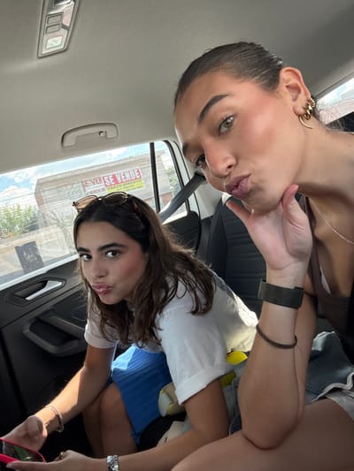 Two young women are depicted inside a vehicle, posing for a selfie. The woman in the foreground, positioned on the right, has dark hair pulled back and light brown eyes. She wears a dark top, a silver chain necklace, a gold-colored ear cuff, a dark green fabric wristband, and a thin brown bracelet on her left wrist. Her left hand is raised to her chin, and she displays a "duck face" expression. The second woman, seated to the left, has dark brown hair with round, brown-rimmed sunglasses perched on her head. She wears a white t-shirt with rolled sleeves and a blue pleated skirt. She holds a pink-cased smartphone in her left hand and wears a silver-toned watch on her left wrist, also exhibiting a "duck face" expression.
The vehicle's interior features a light-colored headliner, a grab handle, and dark-colored door panels. Through the car window, a building is visible under a partly cloudy sky. A large sign with red text on a white background is affixed to the building, stating "SE VENDE" (For Sale) and "TRATO DIRECTO" (Direct Deal), with a partially visible phone number below that begins with "44". This commercial sign in Spanish, coupled with the "44" area code, identifies the location as the city of Pedro Escobedo, Mexico. Green foliage is present outside the vehicle.