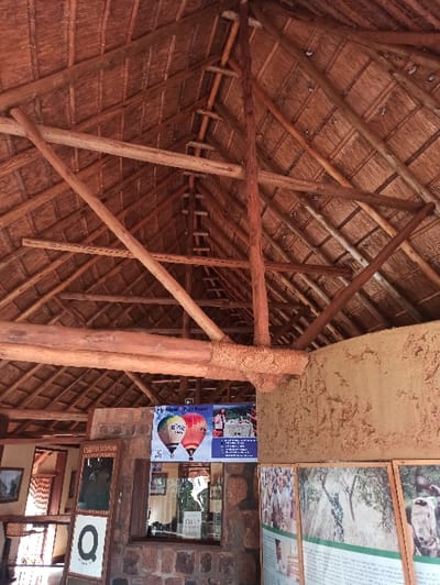 A thatched roof interior is supported by wooden beams and a central column wrapped in woven material. Attached to the adjacent wall are informational posters and framed pictures, suggesting a display or exhibit. The window is visible, offering a glimpse into the surroundings.