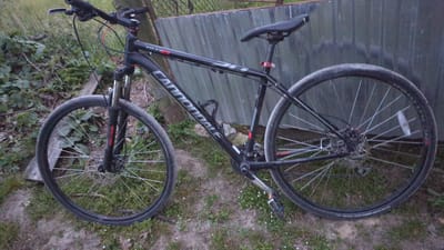 A black Cannondale mountain bike is parked on the side of a dirt path near a corrugated metal fence and some vegetation in Zadebri, Ukraine. The bike is angled to the right, with its front wheel slightly turned. The bike has disc brakes, a water bottle cage mounted to the frame, and a black saddle. The background consists of a wooden fence and some green foliage. The lighting suggests it is either early morning or late evening. No people are present in the image.