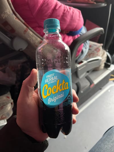 The scene depicts the interior of what appears to be a public transport vehicle, such as a bus or train. A light-skinned hand, partially obscured by a dark sleeve, holds a clear plastic bottle of "Cockta Original" soda. The bottle contains a dark, opaque, carbonated liquid and is topped with a turquoise screw cap. Its label features a turquoise and black design with yellow and white text identifying "FINEST HERBAL EXTRACTS" and "Cockta Original." Embossed text reading "EST. 1953" is visible on the bottle's shoulder.

In the background, to the left of the bottle, a person is partially visible, seated and wearing a bright pink padded jacket. Behind this individual, an upholstered seat with a beige and brown geometric pattern is present. To the right, a dark gray armrest or interior structural component of the vehicle is visible, with a multi-patterned textile item located behind it. A segment of a leg clothed in blue denim is visible in the lower right portion of the frame. The specific location within the city of Cacak, Serbia, cannot be determined from the visual information provided in the image; however, the presence of the "Cockta" brand confirms a regional context within the Balkans.