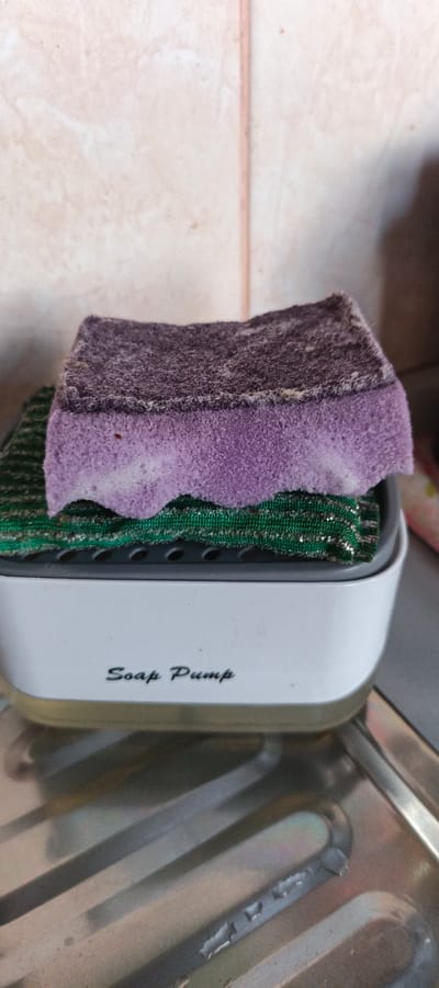 An overhead shot shows a white soap pump dispenser with a green scrubbing pad and a purple sponge on top, resting on a stainless steel sink. The soap pump dispenser has "Soap Pump" written on the front in black script. The background consists of a tiled wall. The sink appears to be in a kitchen or bathroom setting.