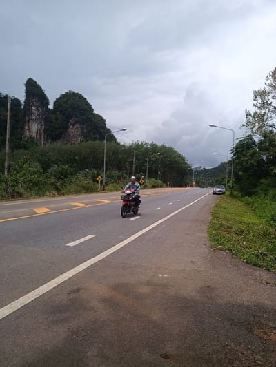 The image depicts a road scene in Khao Khram, Thailand, under a cloudy sky. The asphalt road has a central white line and yellow lines marking the edges of the lanes. A man is riding a small, red motorbike on the road. In the background, there are limestone hills covered with green vegetation. Streetlights line the side of the road, and a dark-colored vehicle is parked on the shoulder. Yellow speed bumps are visible on the road. The shoulder of the road has a grassy verge.